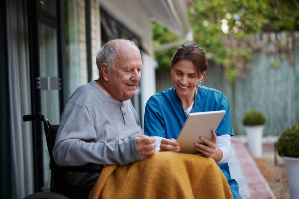 Careful nurse assisting a senior man while using digital tablet on patio in nursing home. Happy nurse and senior patient sitting on wheelchair watching video on digital tablet. Young healthcare professional helping an elderly man use a digital device.